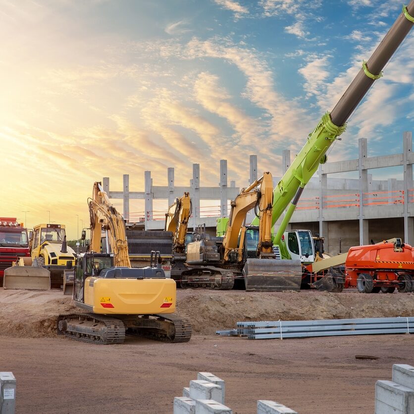 Many different multiclored colorful heavy industrial machinery equipment at construction site parking area against warehouse building city infrastructure development. Commercial vehicles rental sale